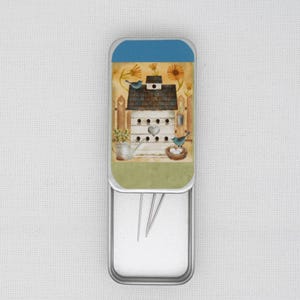 May include: A metal tin with a hinged lid, featuring a colourful illustration of a birdhouse with two bluebirds, a watering can, and flowers. The tin is open, revealing a white interior with three sewing needles.