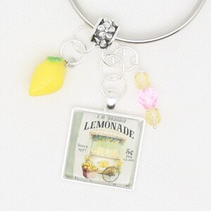 May include: Silver keychain with a charm featuring a vintage lemonade stand illustration. The charm is surrounded by a silver frame and has the text "A.M. Goodman Lemonade" and "Since 1927 5¢ per glass". The keychain also includes a small yellow lemon charm and a pink and yellow bead charm.