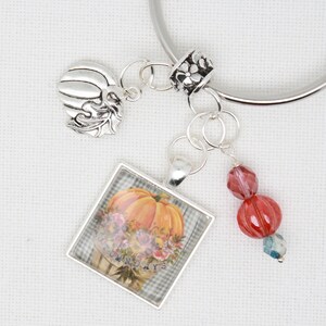 May include: A silver charm bracelet featuring a pumpkin charm, a glass charm with a pumpkin and flowers, and red and clear glass beads. The glass charm has the text "Autumn Days" on it.