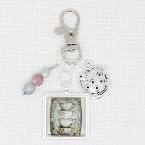 Puede incluir: Llavero plateado con un dije cuadrado que presenta una pila de tazas de té con detalles florales. Un dije de taza de té y una hebra de cuentas pastel cuelgan del llavero. Las tazas tienen una estética vintage.