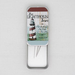 May include: A silver metal tin with a white interior. The lid features a colourful illustration of a lighthouse with the text "The Lighthouse Inn" and "We'll Keep The Light On For You Since 1901".