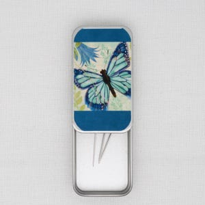 May include: A small metal tin with a blue butterfly design on the lid. The tin is open and contains several sewing needles.