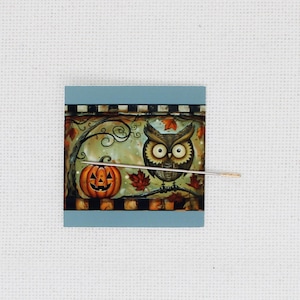 Könnte beinhalten: Ein quadratisches Kunstwerk mit blauem Rand, das ein Halloween-Motiv zeigt. Es zeigt einen geschnitzten Kürbis, eine Eule und Herbstblätter auf einem Ast. Eine Nadel ist in das Kunstwerk eingefügt.