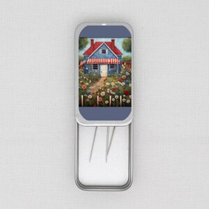 May include: A rectangular, hinged metal tin with a blue lid. The lid displays a watercolour painting of a blue house with a red roof and a white picket fence. The tin contains sewing needles. The house is surrounded by colourful flowers.