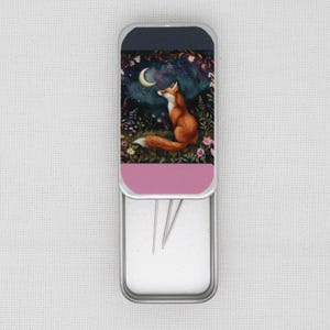 May include: A silver metal needle case featuring a fox design. The case has a pink lid with a fox gazing up at a crescent moon in a starry night sky. The case is open and contains several needles.