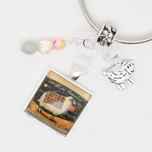 May include: A silver charm bracelet with a square charm featuring a sheep wearing a plaid blanket and holding a jack-o-lantern. The charm is surrounded by a silver frame. The bracelet also includes a silver charm with a flower design and three beads: one green, one clear, and one pink.