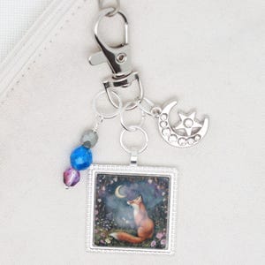 May include: A silver charm keychain with a crescent moon and star charm, a silver frame with a fox looking up at a crescent moon, and a string of purple, blue, and gray beads.