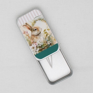 May include: A small metal tin with a hinged lid. The lid features a colourful illustration of a rabbit sitting in a basket of flowers. The tin is open and shows two silver sewing needles inside.