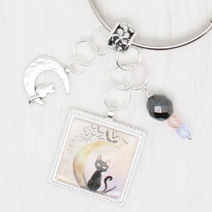 May include: A silver charm bracelet featuring a black cat charm sitting on a crescent moon, a black crystal bead, and a pink and purple crystal bead.