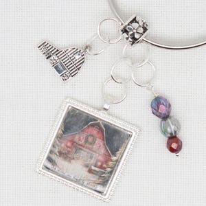 Op de afbeelding: Een zilveren armband met een rode schuurbedel, een zilveren schuurbedel en drie glazen kralen in tinten van paars, grijs en rood. De armband heeft een zilveren ring en een zilveren kraal met een bloemenmotief.