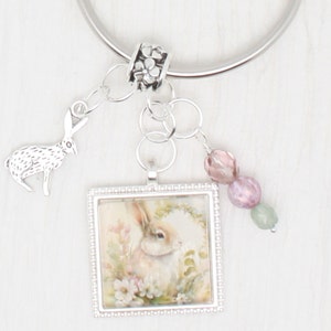 May include: A silver key ring with a charm featuring a picture of a rabbit in a floral setting. The charm is surrounded by a silver frame with a beaded border. The keyring also includes a silver rabbit charm and pink and green beads.
