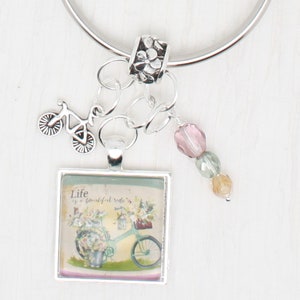 May include: A silver keyring with a charm featuring a bicycle and a square glass charm with a floral design and the text "Life is a beautiful ride". The keyring also has a silver bead with a floral design and three glass beads in pink, green, and yellow.