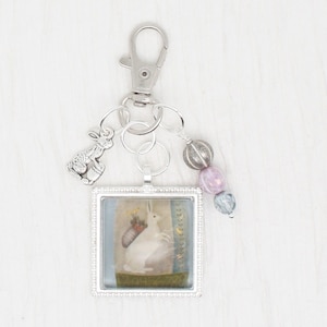 Może przedstawiać: Srebrny brelok do kluczy z kwadratową zawieszką z ilustracją białego królika. Brelok zawiera mały urok królika i ozdobne koraliki w odcieniach szarości, lawendy i niebieskiego. Dołączony jest karabińczyk.
