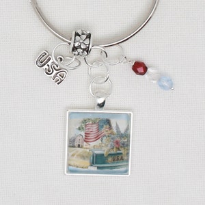 May include: Silver charm bracelet with a square charm featuring an American flag, a red, white and blue crystal charm, and a charm with the word "USA".
