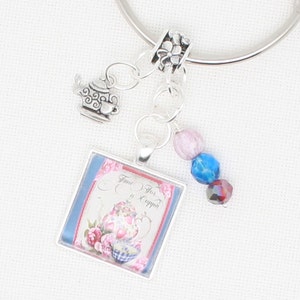 Puede incluir: Pulsera de abalorios plateada con un colgante cuadrado con un diseño floral de tetera y el texto "Time For a Cuppa". La pulsera incluye un charm de tetera y un grupo de cuentas rosas, azules e iridiscentes. Fondo claro.