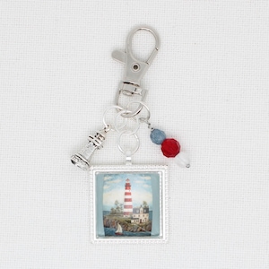 May include: A silver-coloured keychain with a nautical theme. It features a square pendant with a lighthouse scene, a small lighthouse charm, and red, white, and blue beads. The keychain has a lobster claw clasp.