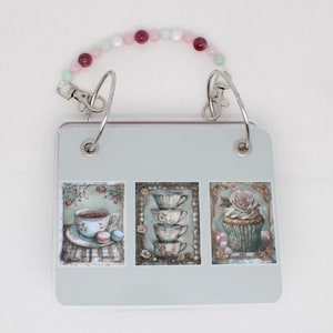 May include: A gray photo album with a beaded handle. The album has three images on the front: a teacup with macarons, a stack of teacups, and a cupcake with a rose.