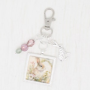 May include: A silver charm keychain with a square picture frame featuring a rabbit in a floral setting. The keychain also includes a small rabbit charm and pink and green beads.