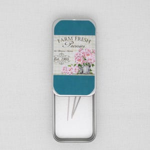 May include: A silver metal tin with a teal lid. The lid has a floral design with pink peonies and the text "FARM FRESH Peonies Buds + Blooms + Stems Est. 1905". The tin is open and contains three silver needles.