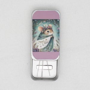 May include: A small metal tin with a pink lid and a floral design featuring a mouse wearing a crown and a cloak. The tin is open and contains three sewing needles.