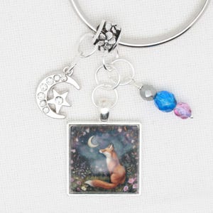 May include: A silver charm bracelet featuring a fox charm, a crescent moon charm, and a silver bead with floral embellishments. The fox charm depicts a fox gazing upwards at a crescent moon in a woodland scene. The bracelet also includes a blue and pink crystal bead.