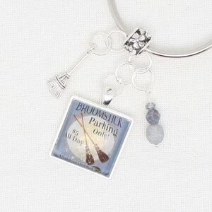 May include: A silver charm with a square charm that says "Broomstick Parking Only! £5 All Day" with two broomsticks crossed in front of a moon. The charm is attached to a silver chain with a silver flower bead and two grey beads.