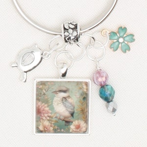 May include: Silver charm bracelet with a bird pendant. The pendant features a square image of a bird perched on a branch surrounded by flowers. Additional charms include a teal flower, beads, and a turtle charm.