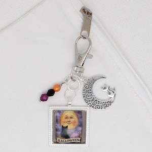 以下が含まれることがあります： ハロウィーンをテーマにしたキーホルダー。シルバーの留め金が付いています。笑顔の月と黒猫の正方形ペンダント、猫のシルエットの三日月チャーム、紫、黒、オレンジ色のビーズが付いたアクセントが特徴です。