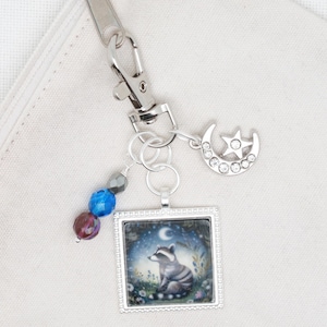 May include: A silver charm keychain with a moon and star charm, a silver frame with a raccoon illustration, and blue and purple glass beads.