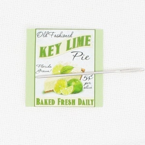 May include: A vintage-style sign advertising Key Lime Pie, with a green background and white lettering. The sign says "Old Fashioned Key Lime Pie", "Florida Grown!" and "Baked Fresh Daily". There is a picture of a key lime pie with a slice cut out, and a needle is placed on top of the sign.