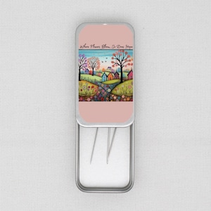 May include: A small, rectangular metal needle case with a pink lid featuring a colourful landscape painting. The painting depicts a village scene with rolling hills, trees, and flowers. The text "Where Flowers Bloom, So Does Hope" is printed on the lid.