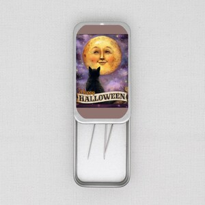May include: A silver needle case with a Halloween-themed design. The artwork features a smiling moon, a black cat, and the words "Happy Halloween" on a banner. The case is open, revealing several needles.
