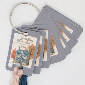 May include: A set of gray rectangular cards held together by a silver metal ring. Each card features a printed illustration of a stone house surrounded by autumn trees and the text, To everything there is a season and a time to every purpose under the heaven. Two strands of embroidery floss in light pink and dark blue are attached to the bottom.