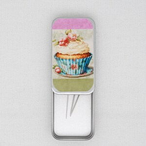 May include: An open, silver metal tin with sewing needles inside. The lid is decorated with a watercolour illustration of a cupcake with pink flowers, a blue and white striped wrapper, and a pink and green background.