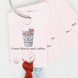 May include: A pink card with a coffee cup illustration and the text "Cross Stitch and Coffee". The card has holes for thread and is part of a set of cards with a metal ring.