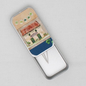 May include: A silver metal tin with a hinged lid. The lid features a colourful illustration of a small house with a sign that reads "Sean and Liam's". The house is decorated with green shamrocks and a string of green shamrock flags.