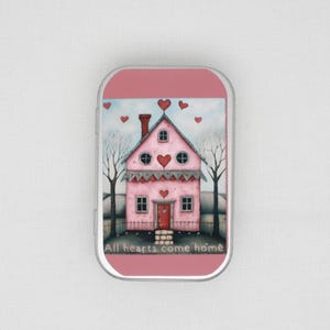 Puede incluir: Una caja de metal rosa con una ilustración caprichosa de una casa rosa con una puerta roja, ventanas en forma de corazón y una chimenea. La casa está situada sobre un fondo de cielo azul con corazones y árboles desnudos. El texto "All hearts come home" está en la parte inferior.