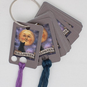 May include: A set of gray Halloween-themed tags attached to a silver ring. Each tag features a smiling moon, a black cat, and the words "Happy Halloween." Purple and teal tassels hang from the tags.