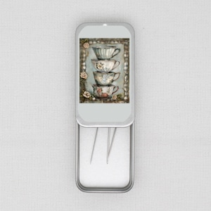 May include: A silver metal tin with a white background and a colorful image of four stacked teacups with floral designs. The tin is open and reveals a white interior with three silver needles.