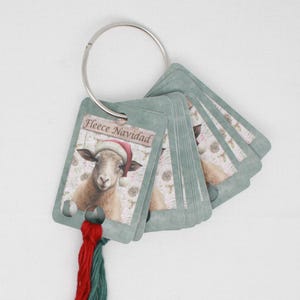 Puede incluir: Un conjunto de marcadores de punto de Navidad de color verde y blanco con una oveja que lleva un gorro de Papá Noel en la parte delantera. Los marcadores están unidos a un anillo plateado y tienen colas de hilo rojo y verde.