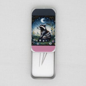 May include: A silver metal needle case with a pink lid featuring a raccoon illustration. The raccoon is sitting in a field of flowers under a crescent moon and stars. The case is open and contains several needles.