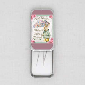 May include: A silver metal tin with a pink lid featuring a cartoon bunny holding an umbrella with the text "April Showers Bring May Flowers". The tin is open and contains sewing needles.