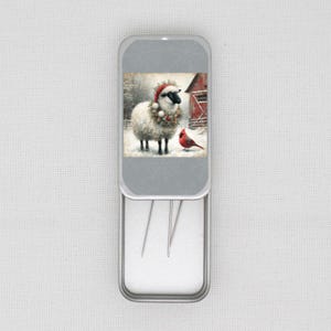 May include: A silver needle case with a winter scene featuring a sheep wearing a Santa hat and a cardinal bird. The case has a grey snowflake pattern on the lid and contains several needles.