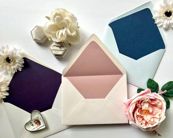 May include: An overhead shot of three open envelopes with colored interiors: navy, light blue, and blush pink. The envelopes are surrounded by white flowers, a pink rose, and a small heart-shaped dish with a stamp. A ring box and a ring are also visible.