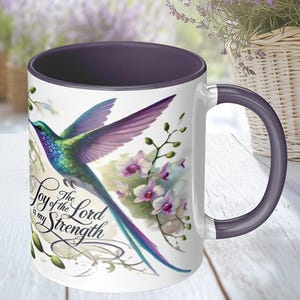 Puede incluir: Taza de cerámica blanca con borde y asa morados. La taza presenta un colibrí colorido con una cola larga y el texto "The Joy of the Lord is my Strength".