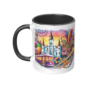 May include: White ceramic mug with a black rim. The mug features a colorful illustration of a New Orleans street scene with a streetcar, a church, a trumpet, and a Mardi Gras mask. The background is a watercolor sunset.