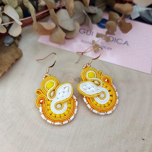 May include: A pair of yellow and white soutache earrings with gold-tone ear hooks. The earrings feature an abstract design with detailed stitching and beadwork. The earrings are presented on a wooden surface, showcasing the craftsmanship.