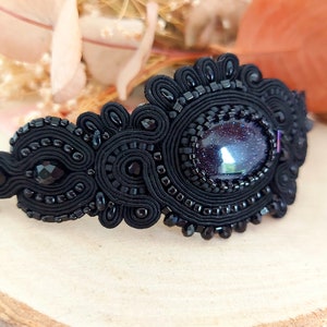 May include: Black beaded bracelet featuring a large, oval-shaped, iridescent blue gemstone at its centre. The bracelet is crafted with black thread and beads, showcasing a delicate and intricate design.
