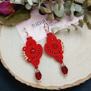 Op de afbeelding: Rode en gouden oorbellen met een hangende rode kristal. De oorbellen zijn gemaakt van rood garen en hebben een delicaat, ingewikkeld ontwerp.