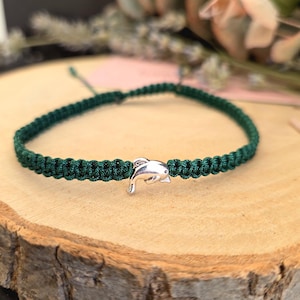 May include: A dark green macrame bracelet with a silver dolphin charm. The bracelet is knotted and rests on a wooden surface. The dolphin charm is detailed, adding a nautical touch to the accessory.